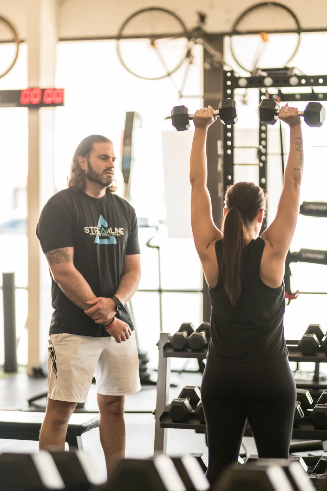 A coach standing beside an athlete pressing dumbbells overhead at Streamline Performance Physical Therapy in Phoenix, AZ